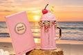 Pink Island Breeze Armaf Breeze spray packaging box and matching pink whipped-cola-style mug with gold trim, a cherry topper, and a striped straw, displayed on rocks at a beach sunset.