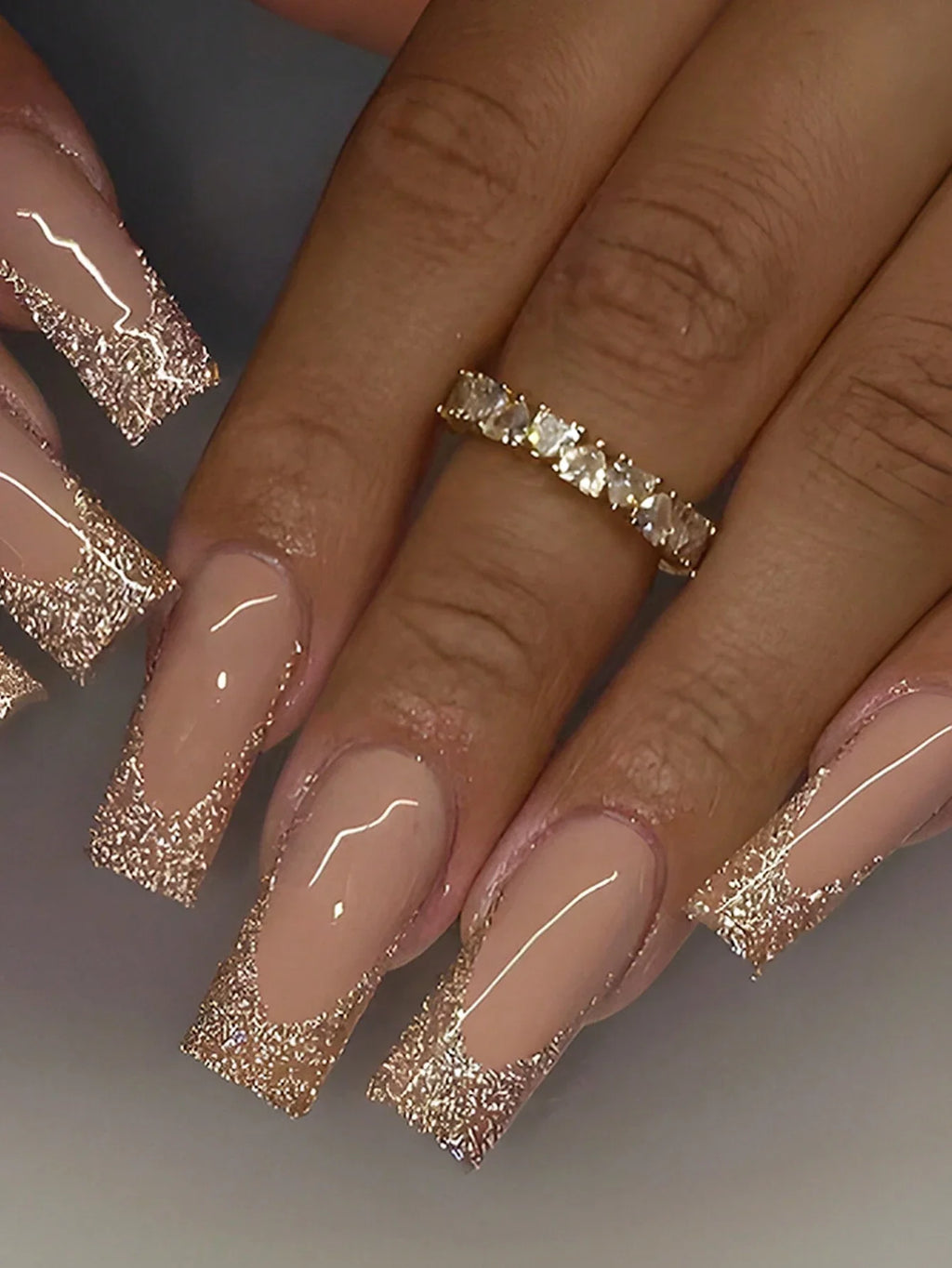 Close-up of a hand with beige nails and gold glittery tips, wearing a gold ring.