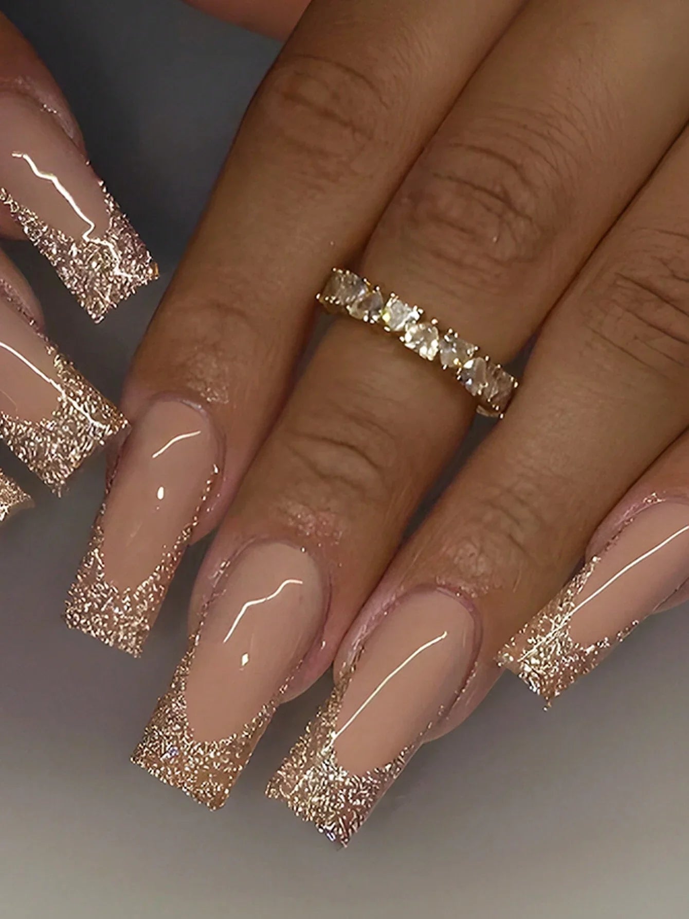 Close-up of a hand with beige nails and gold glittery tips, wearing a gold ring.